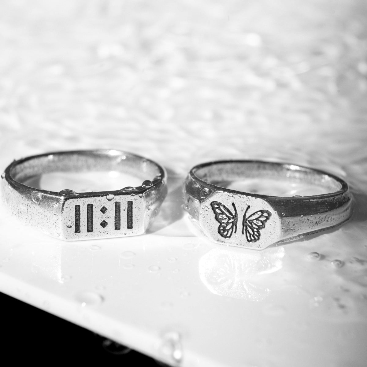 A black and white shot of the sterling silver Momento Ring and another signet ring from Australian jewellery brand Billie Jo, resting on a wet surface.