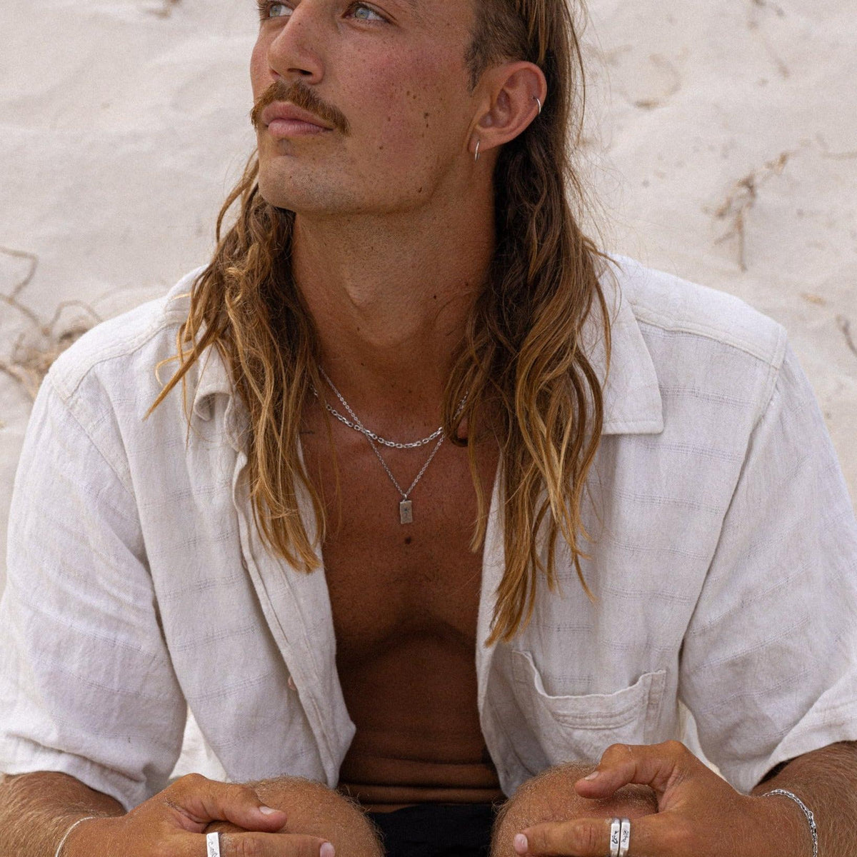 A male model with long hair and a moustache, wearing an open white shirt, showcases the Billie Jo KOI I Ring and other layered jewellery on a sandy beach.