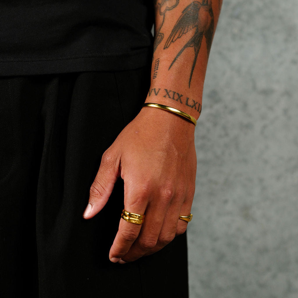 A man's tattooed arm wearing the 18k gold vermeil Drifter and Union set by Billie Jo. The sleek gold cuff and signet ring are styled with a black tee for a modern, masculine look.