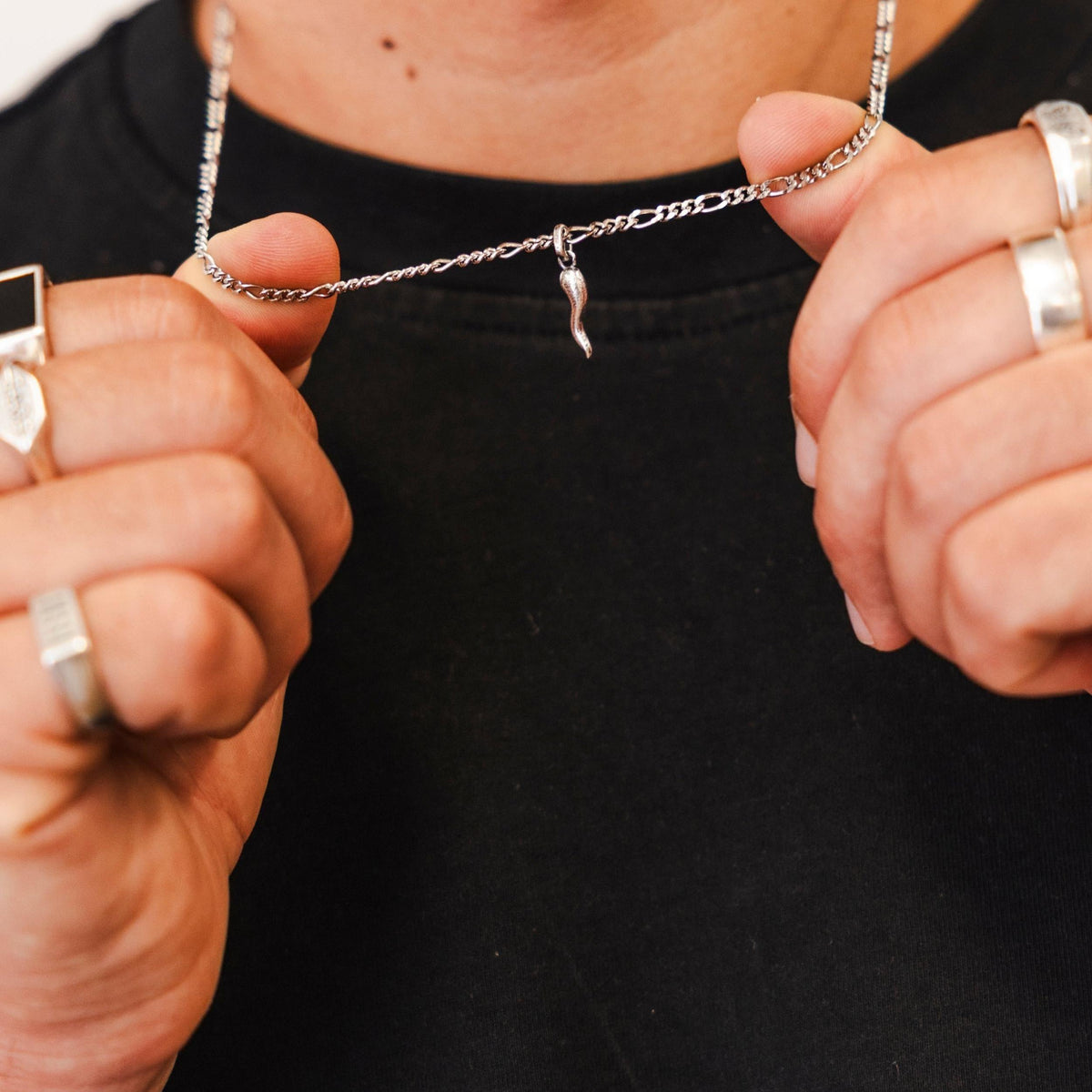 A male model holds the chain of the Billie Jo Chilli Pendant, showcasing the silver horn charm against a black t-shirt. He also wears several silver rings.