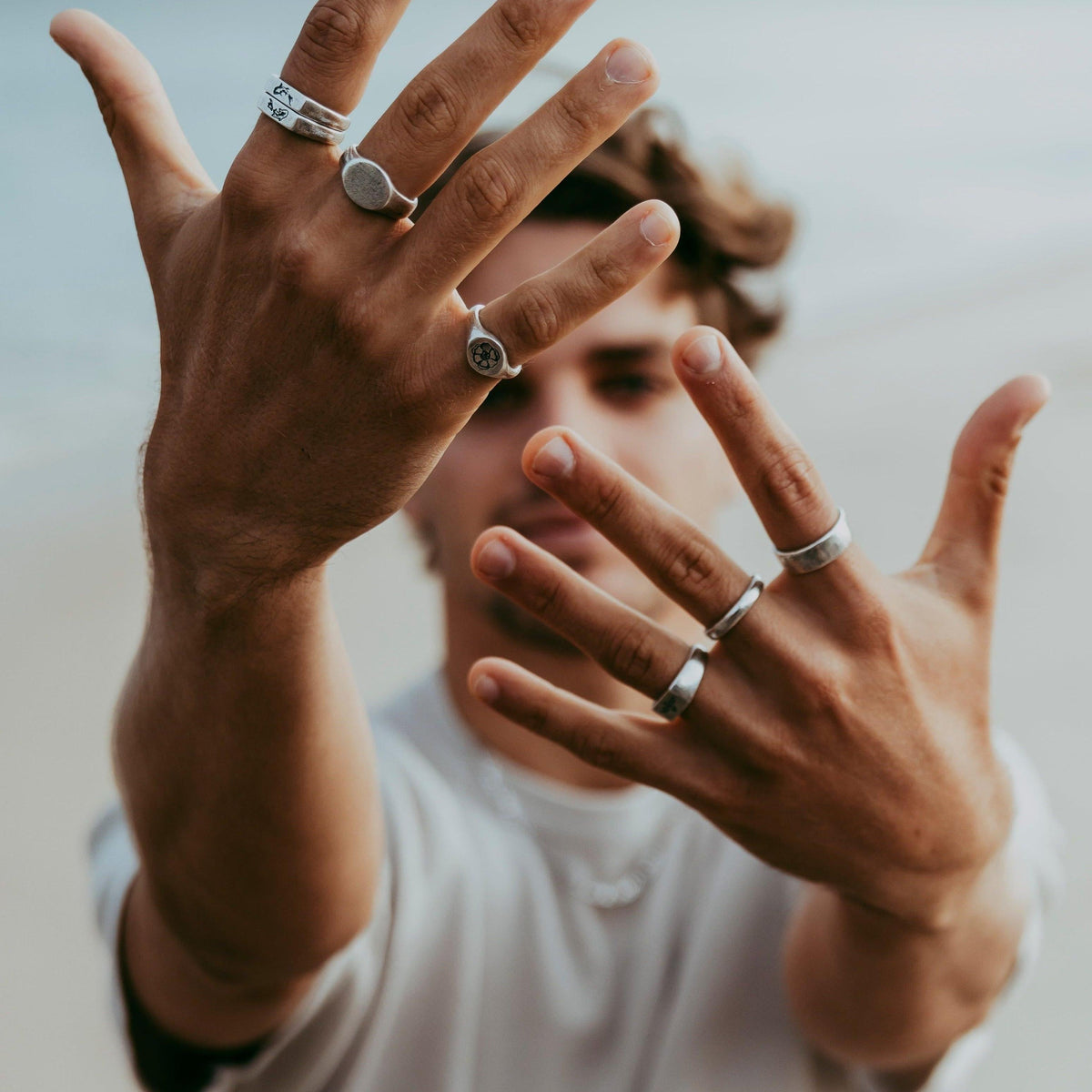 A male model with wavy hair holds his hands up to the camera, showcasing a collection of sterling silver rings from Billie Jo, including the KOI I Ring.