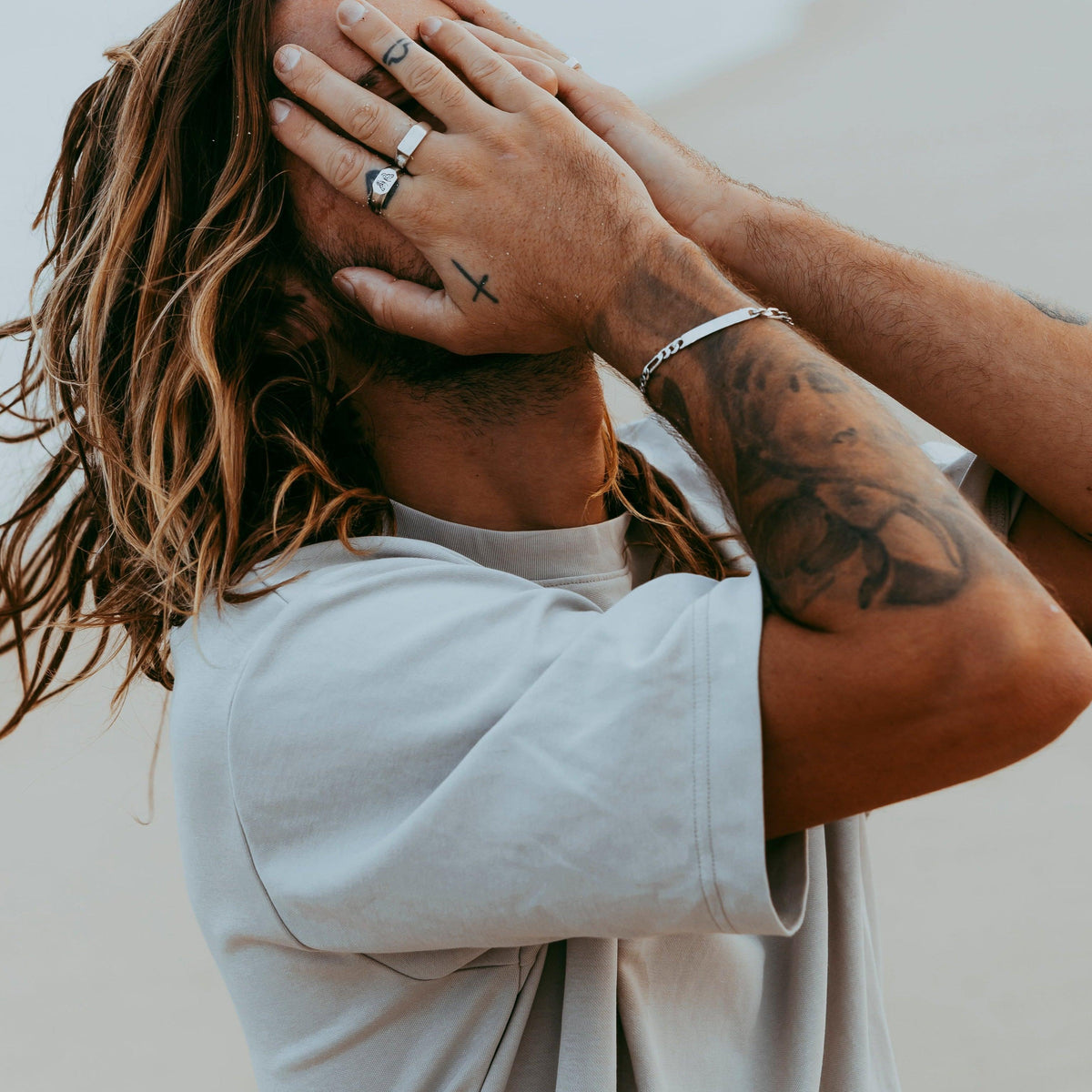 A male model with long, sun-kissed hair and tattooed arms wears the Billie Jo Genesis Ring and other jewellery. A modern, masculine look for the Australian man.