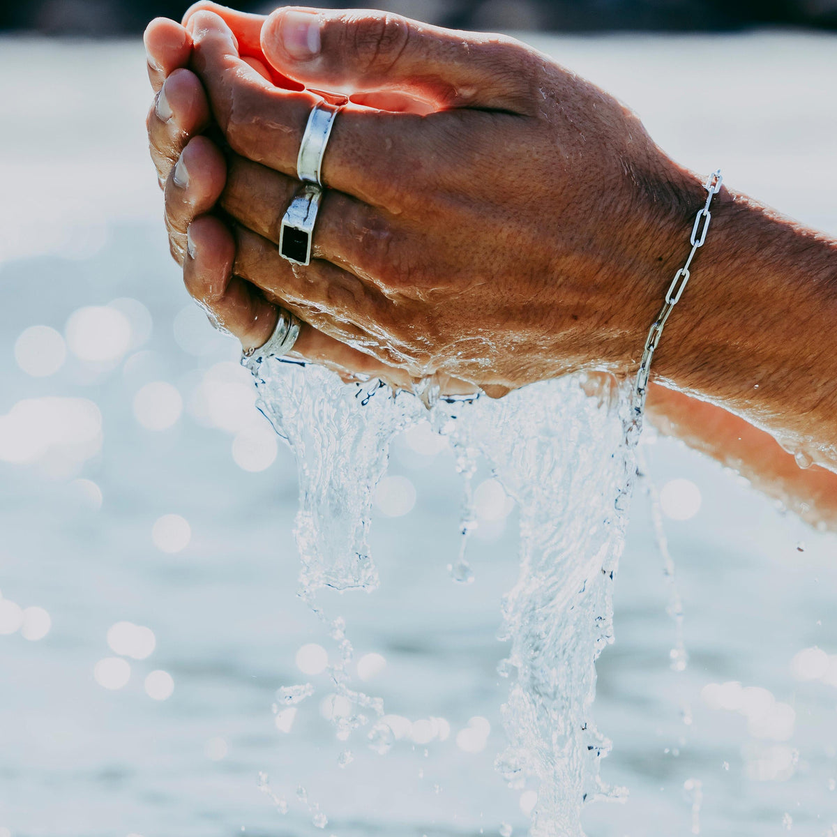 A man's hands cupping water, wearing the sterling silver Link Bracelet and multiple rings from Billie Jo. The water cascades from his hands, with the sparkling ocean in the background. This is a detailed lifestyle shot from the Australian jewellery brand's campaign.