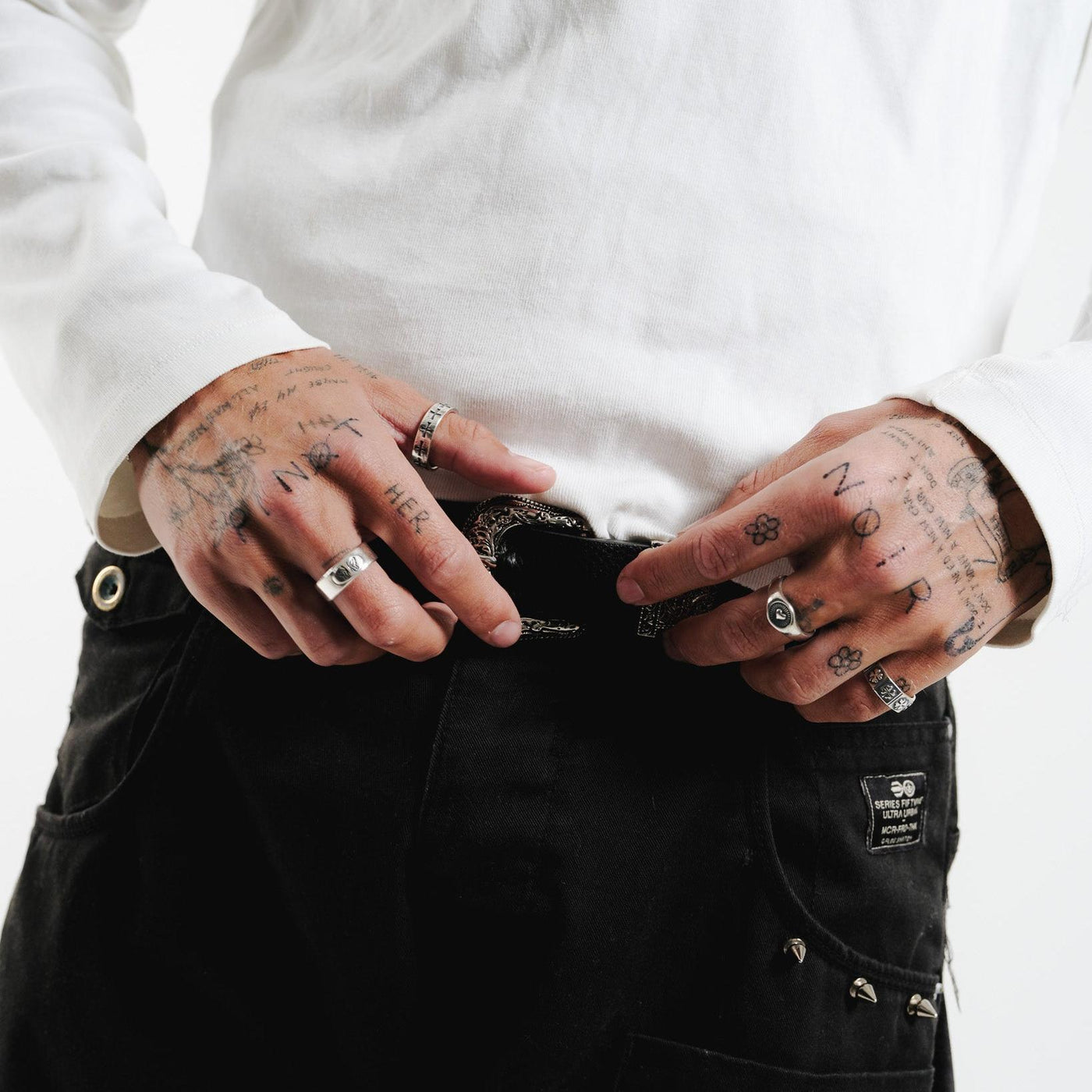 A close-up of a man's hands with tattoos, wearing the Billie Jo Sanctum Ring and other silver jewellery. He is adjusting a decorative belt buckle.