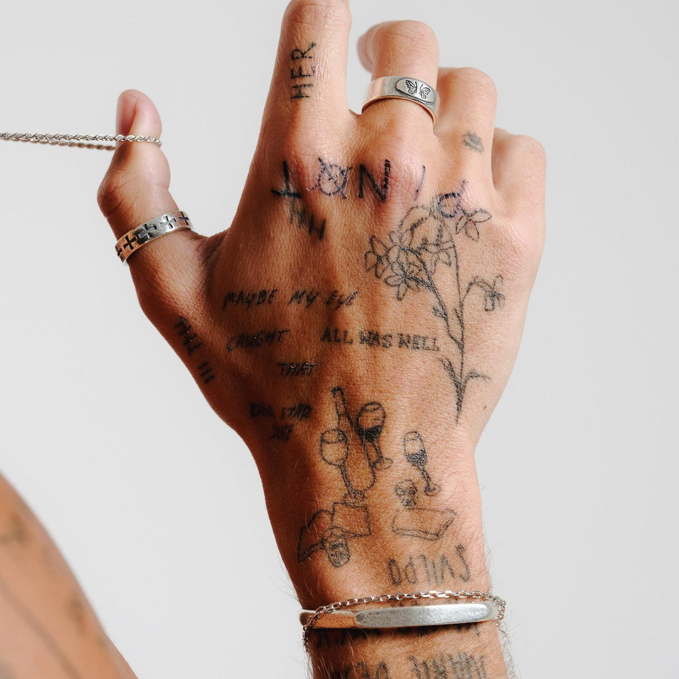 A tattooed hand, adorned with the Billie Jo Sanctum Ring and a silver bracelet, delicately holds a silver chain. An artistic shot showcasing men's jewellery.