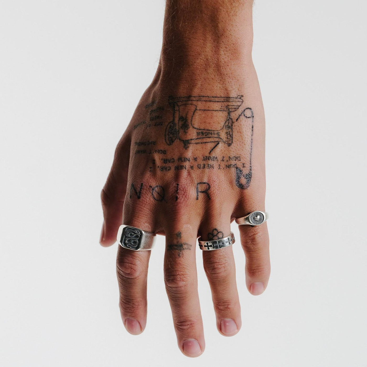 A man's tattooed hand wearing a stack of silver rings, including the Billie Jo Sanctum Ring. This image highlights the ring's versatile, stackable design.