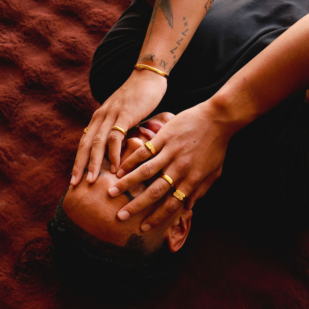 An artistic shot of a man lying down, with his hands covering his face, adorned with various gold rings from Billie Jo, including the Stacker Ring. The mood is intimate and stylish.