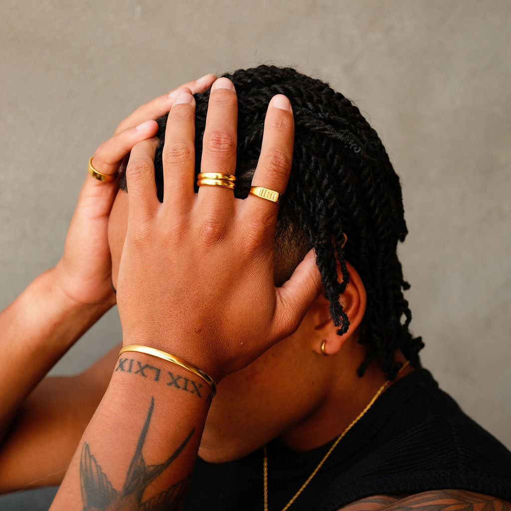 A man with braided hair wearing multiple gold rings, including the Billie Jo Momento Ring, showcasing a modern, layered men's jewellery style.