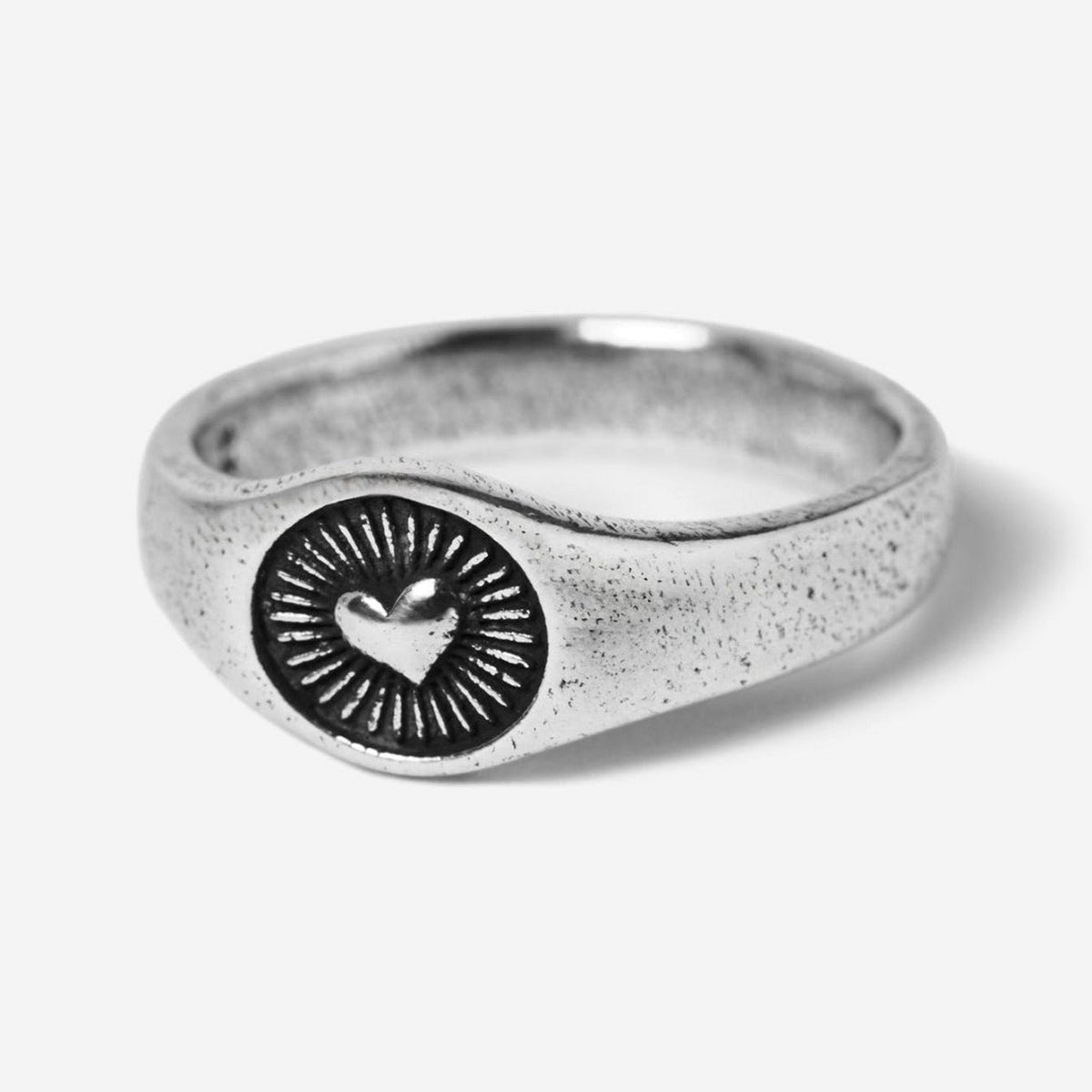 A 45-degree product shot of the sterling silver Vita Ring by Billie Jo. The signet-style ring features a central heart emblem with radiating sunbeam engravings, showcasing its textured, oxidised finish.