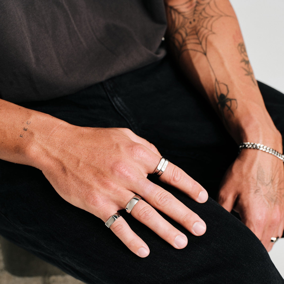 Man wearing silver finish stainless rings, seated, hand resting on his thigh, with a steel Cuban bracelet on the other wrist