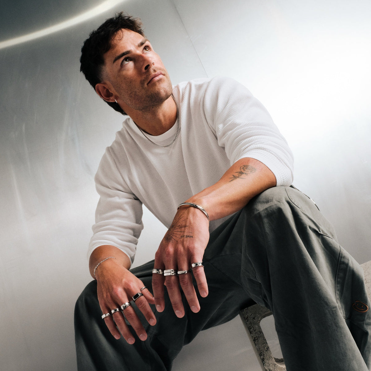 Man wearing silver finish stainless rings, seated, looking up, wrists on his knees, white waffle long sleeve, olive pants