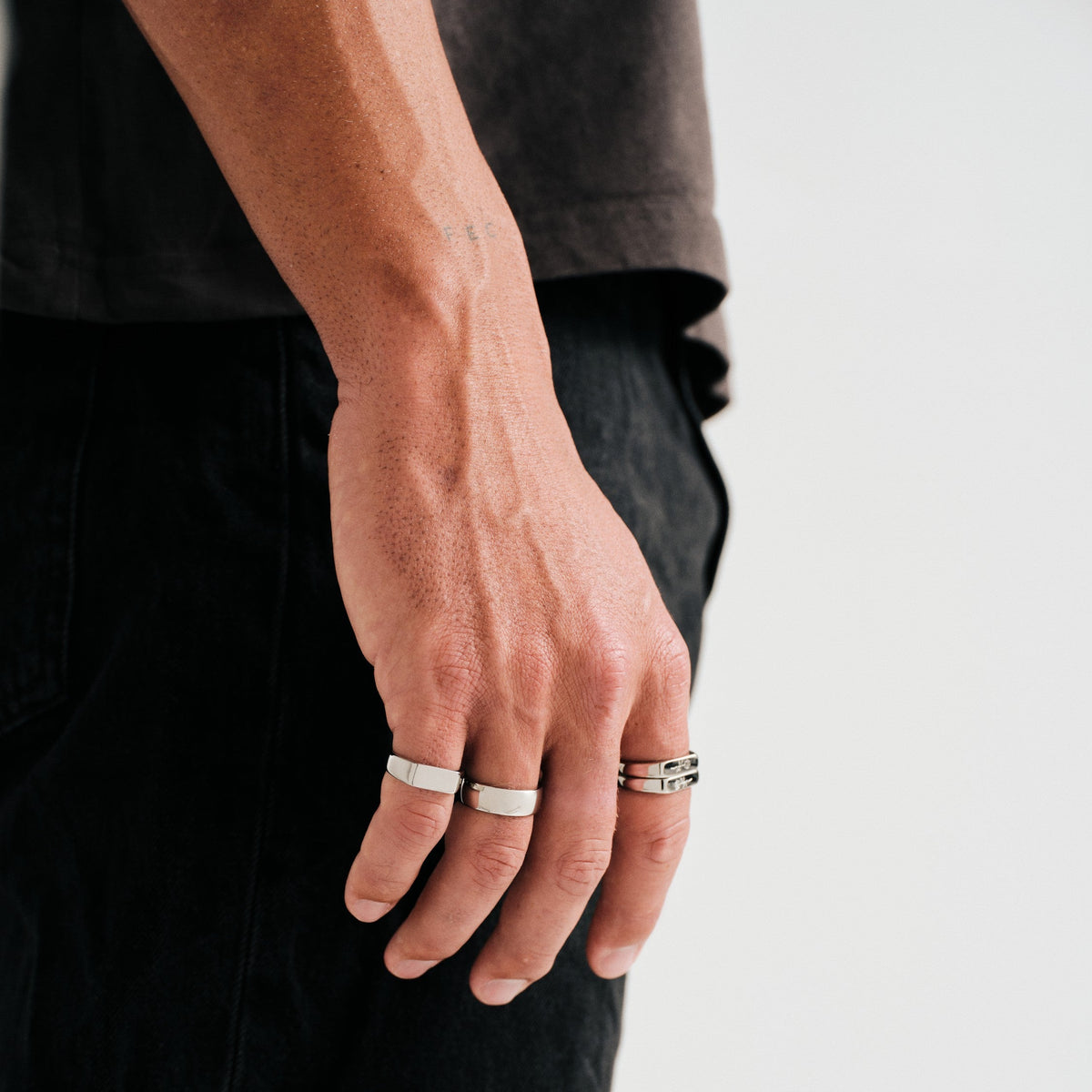 Man wearing silver finish stainless rings, hand hanging by his side, brown tee, black jeans