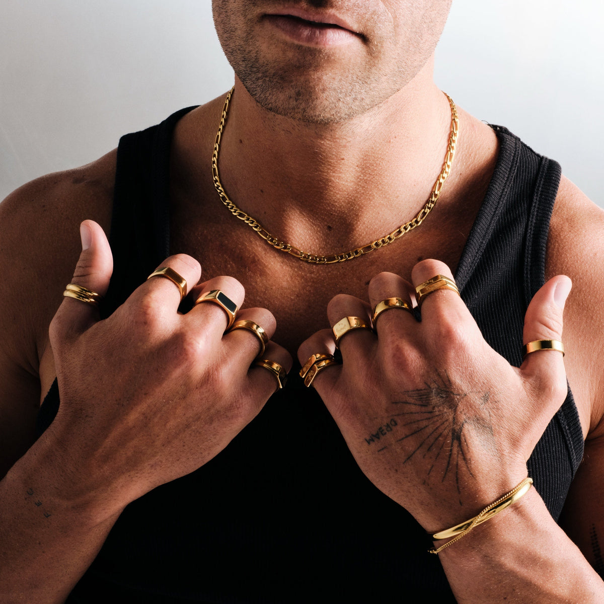 Man wearing gold stainless rings, hands on his chest, with a gold Figaro chain, gold cuff and slim bracelet, black singlet