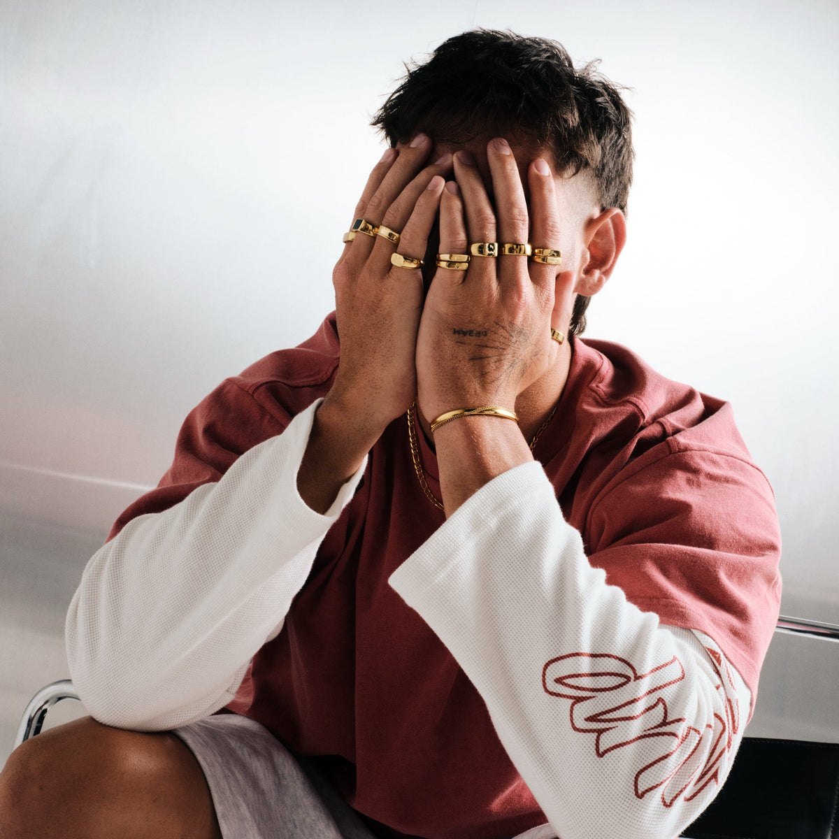 Man wearing gold stainless rings, seated, hands over his face, with a gold chain necklace, gold cuff