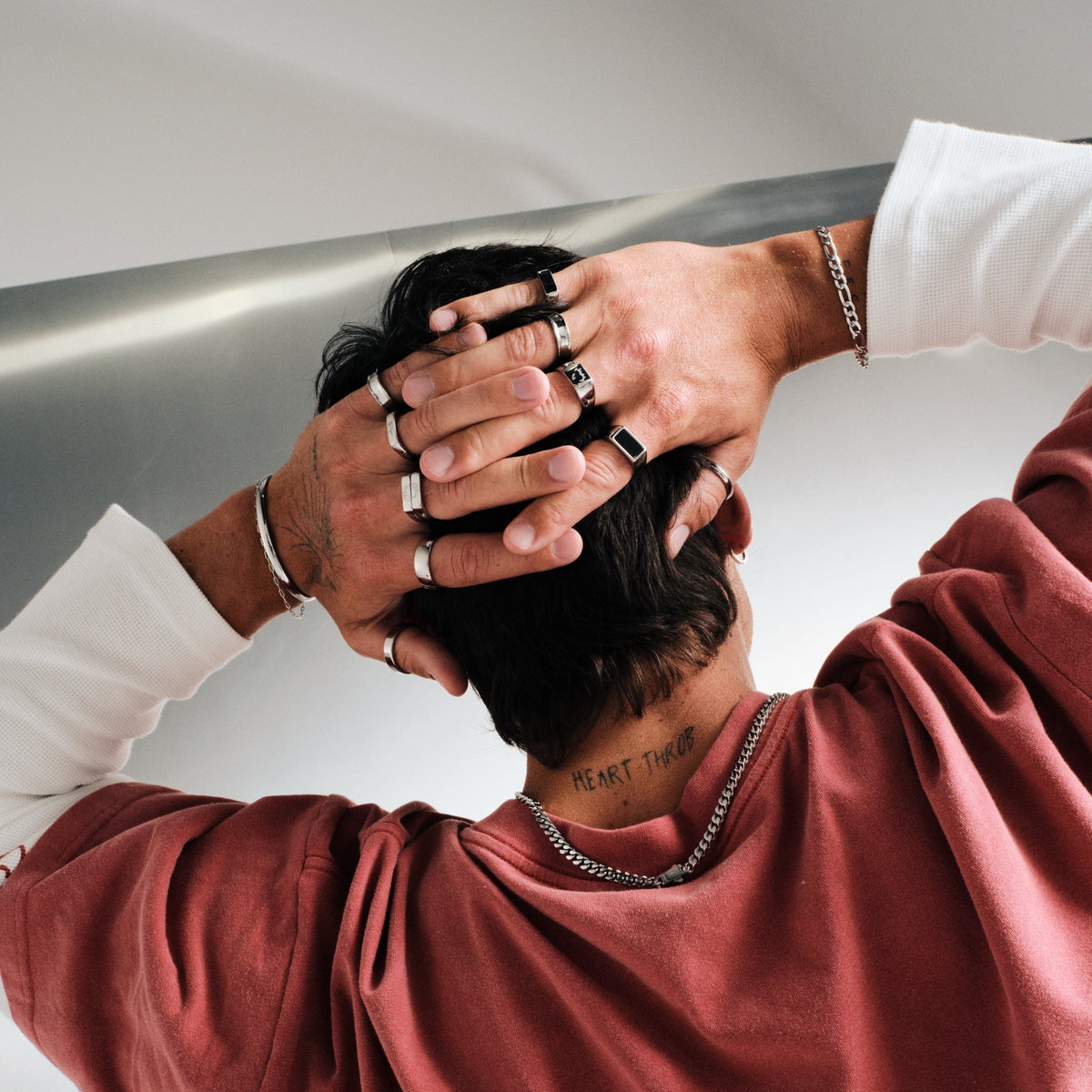 Man wearing silver finish stainless rings, hands behind his head, back to camera, red tee over white long sleeve