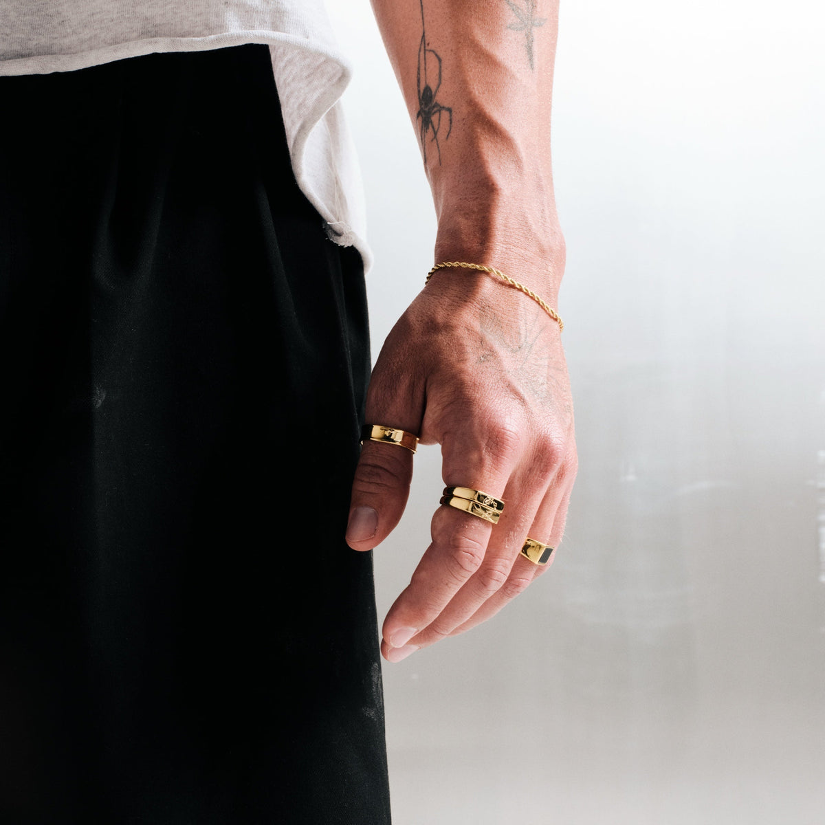 Man wearing gold stainless rings, hand by his side, with a gold rope bracelet, white tee, black pants