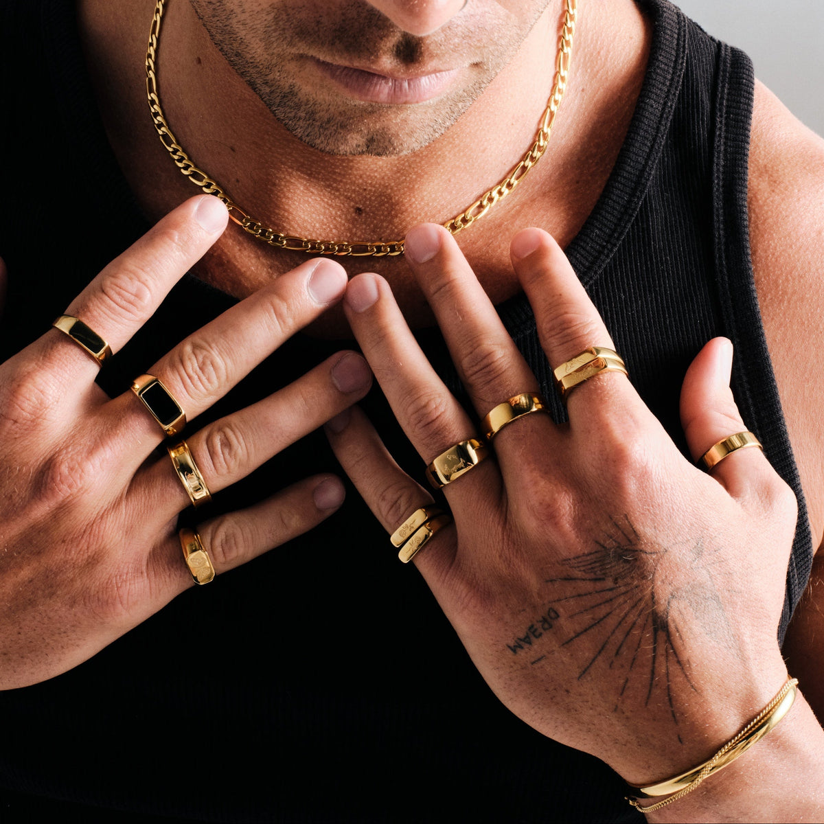 Man wearing gold stainless rings, hands on his chest, with a gold Figaro chain necklace, gold bangle and chain bracelet