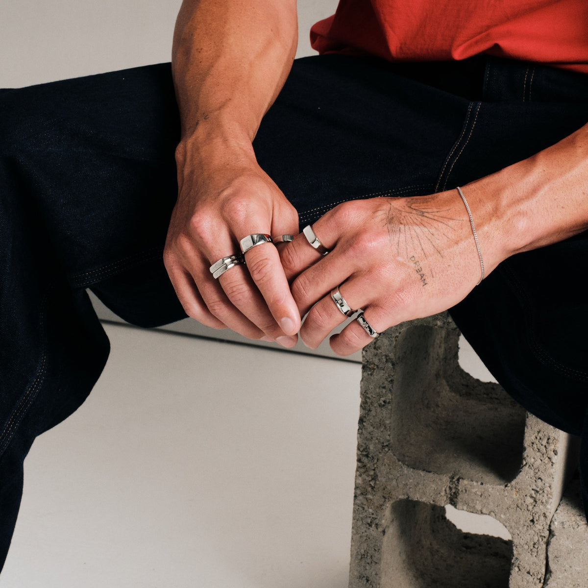 Man wearing silver finish stainless rings, seated on a concrete block, hands clasped, with a fine steel chain bracelet