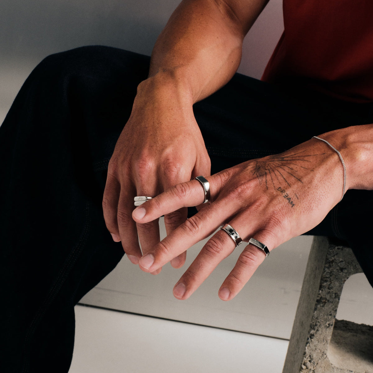 Man wearing silver finish stainless rings, seated on concrete block, one hand touching a ring on the other