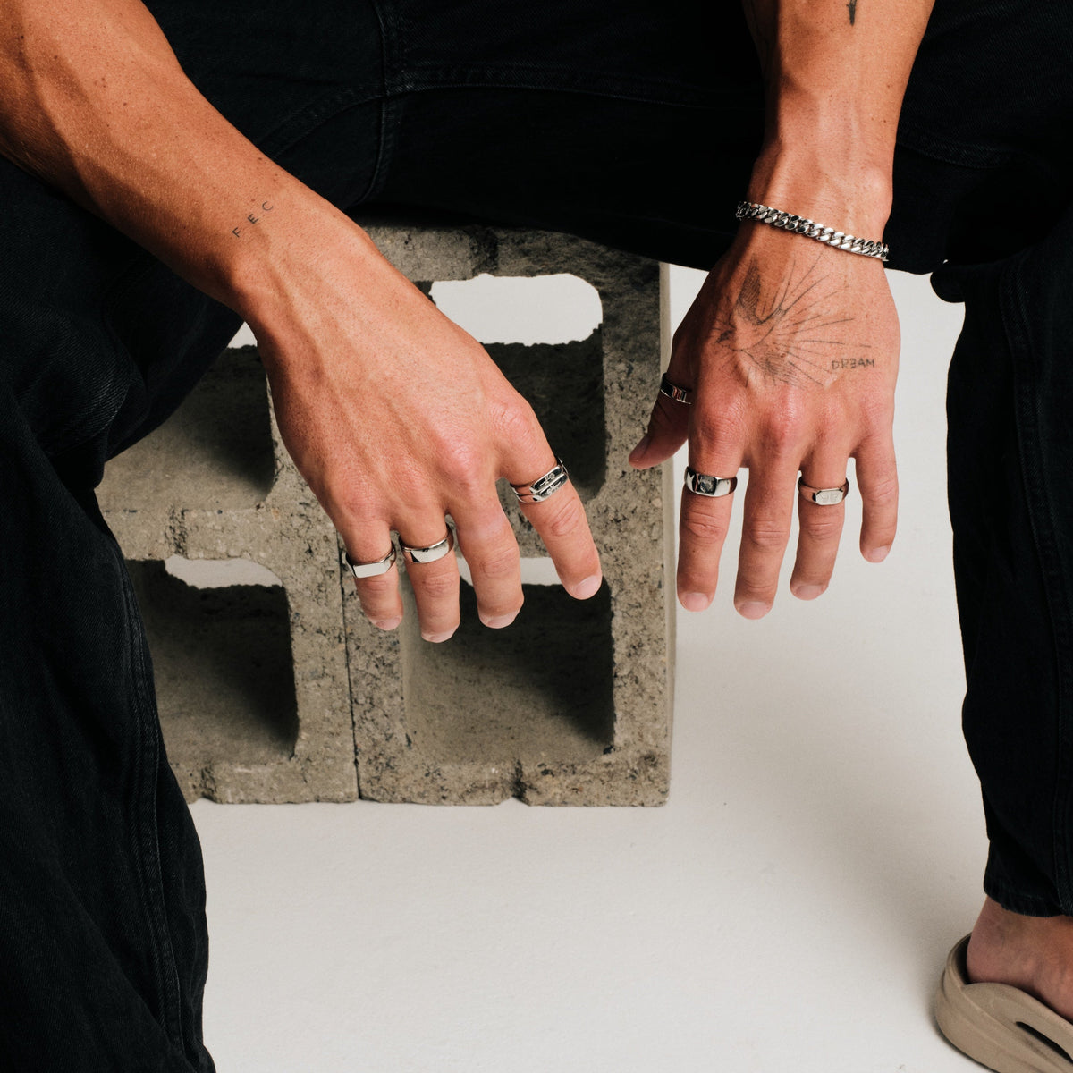 Man wearing silver finish stainless rings, crouched on concrete block, hands hanging down, black jeans, sandals