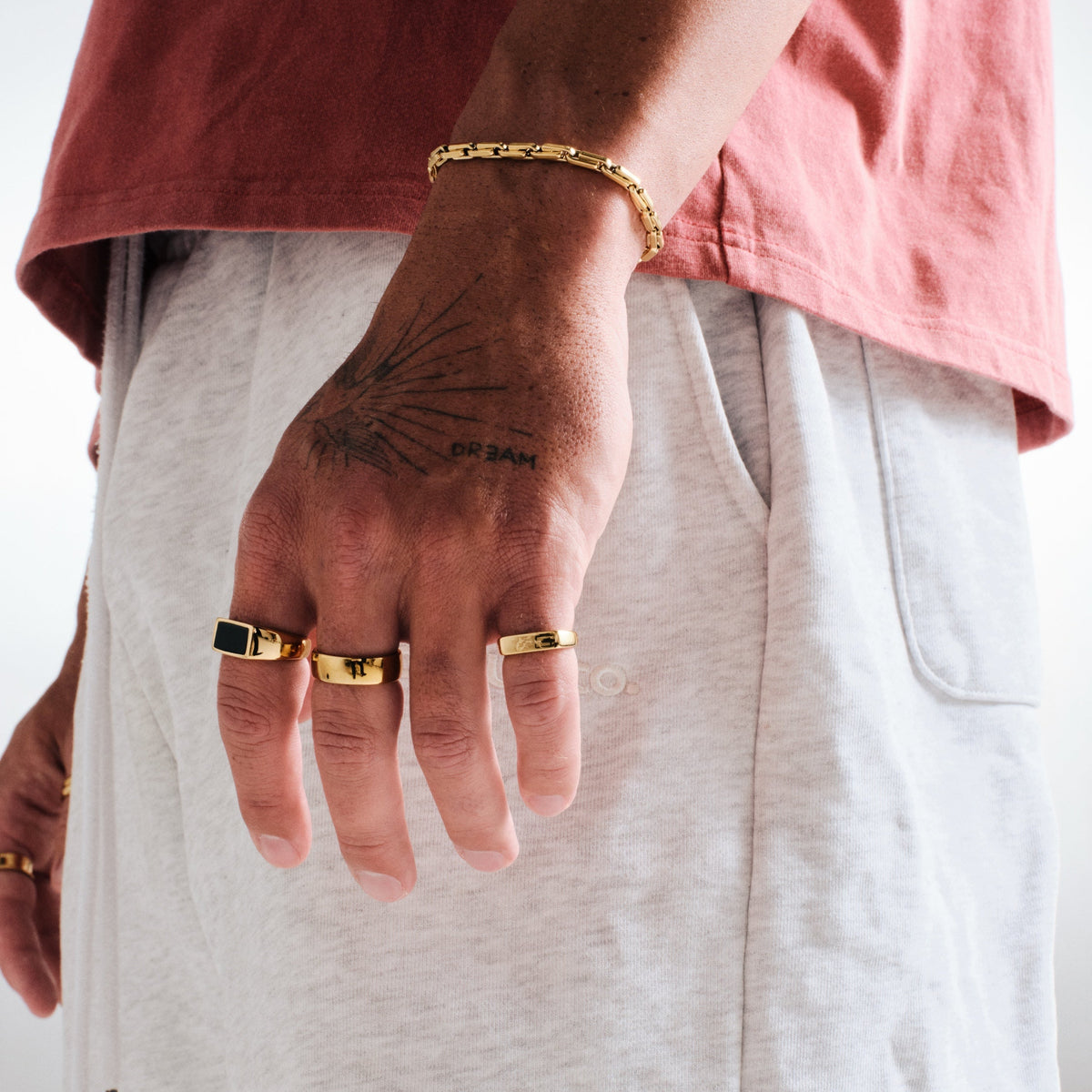 Man wearing gold stainless rings, hand relaxed at his side, with a gold box link chain bracelet, red tee, grey track pants