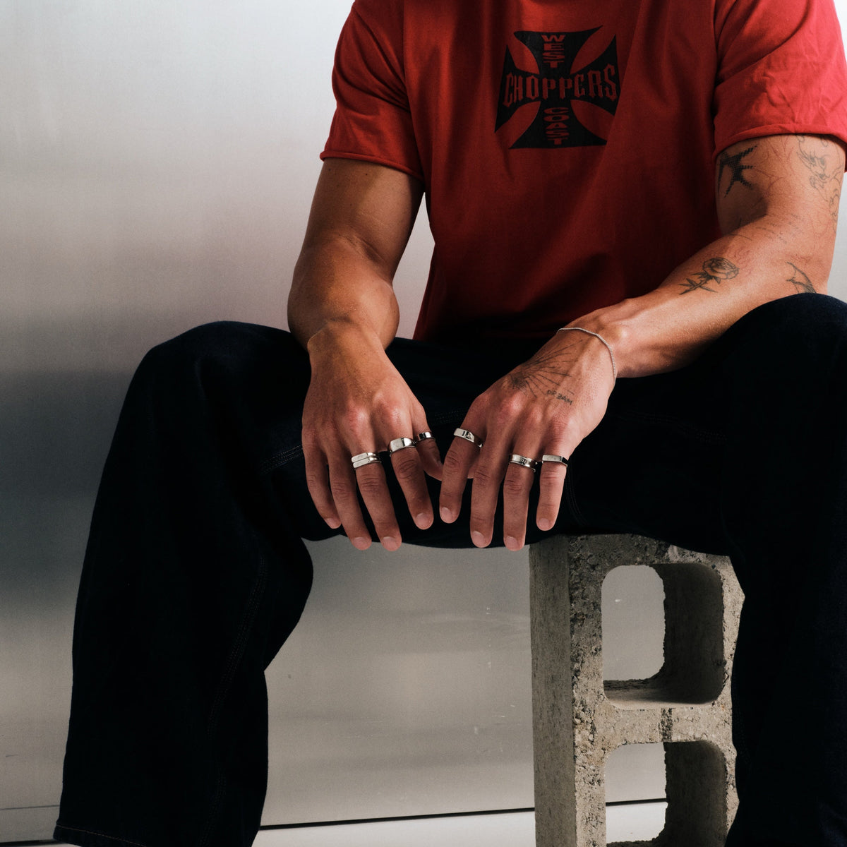 Man wearing silver finish stainless rings, seated on a concrete block, hands on knees, with a bangle on left wrist
