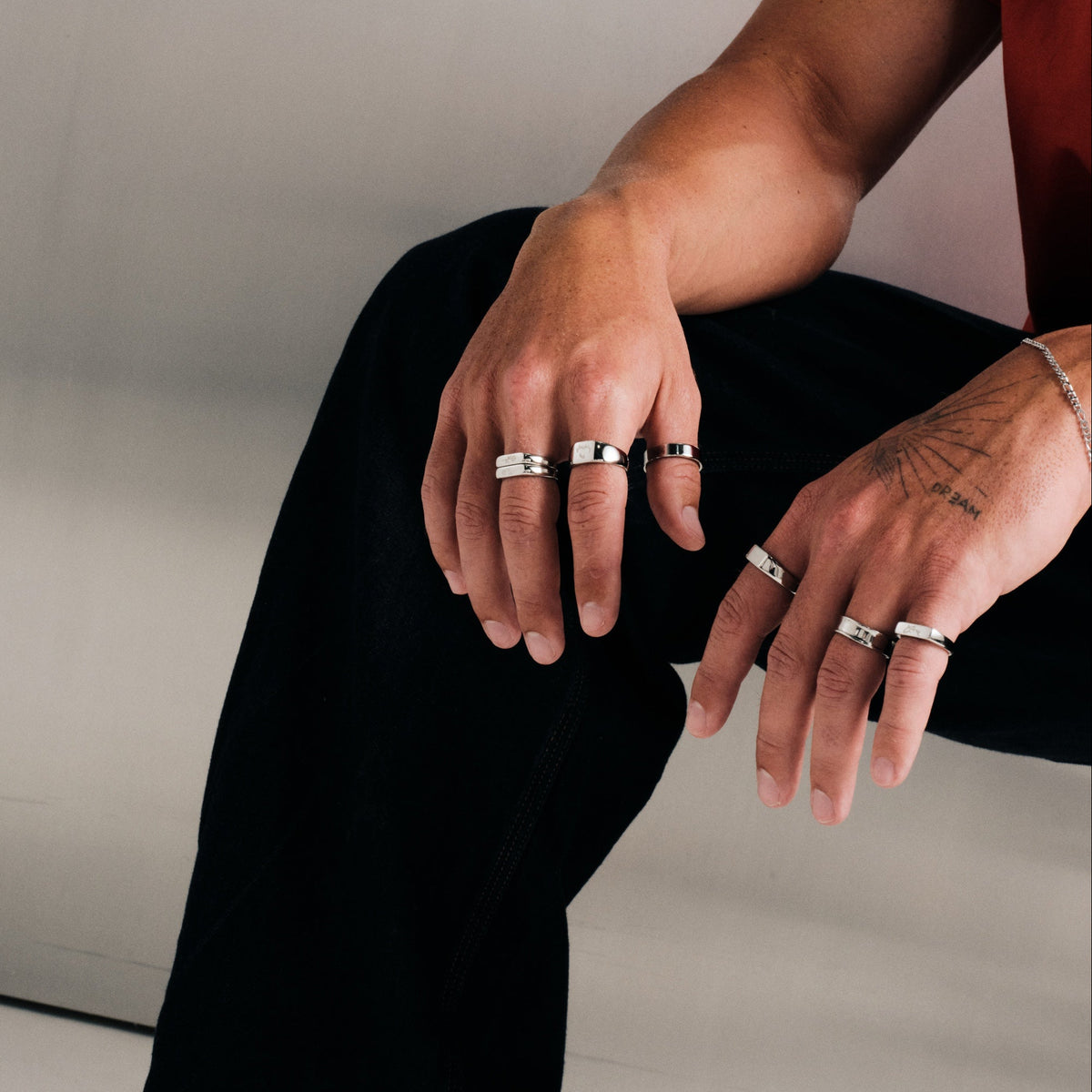 Man wearing silver finish stainless rings, seated, hands hanging over his knee, with a slim chain bracelet