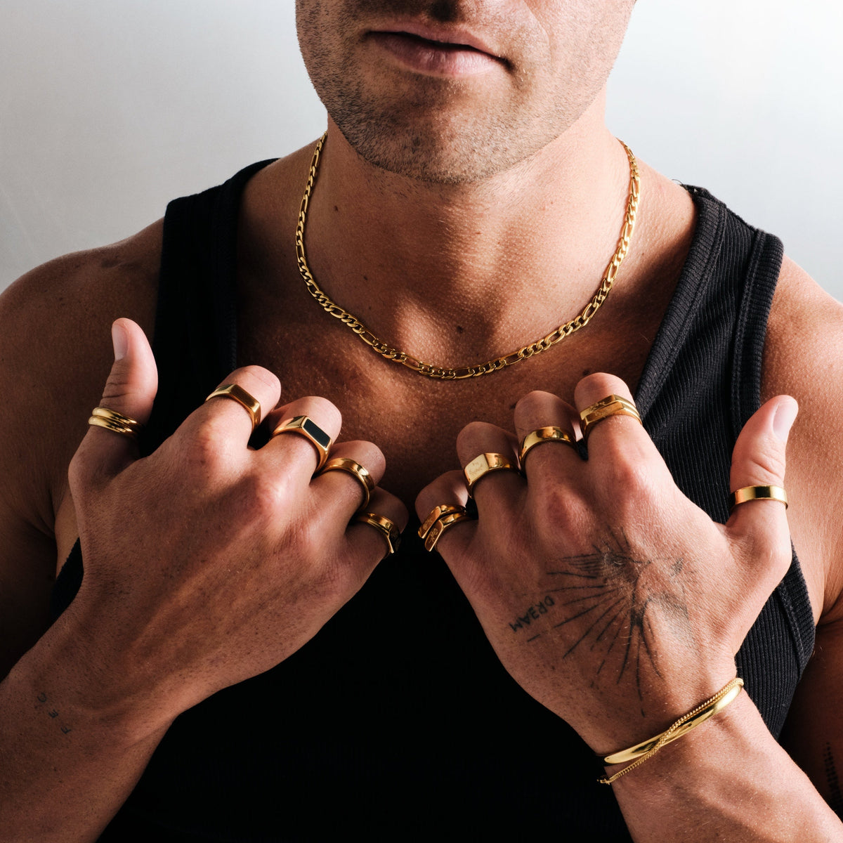 Man wearing gold stainless rings, hands on his chest, with a gold Figaro chain necklace, gold bangle and chain bracelet
