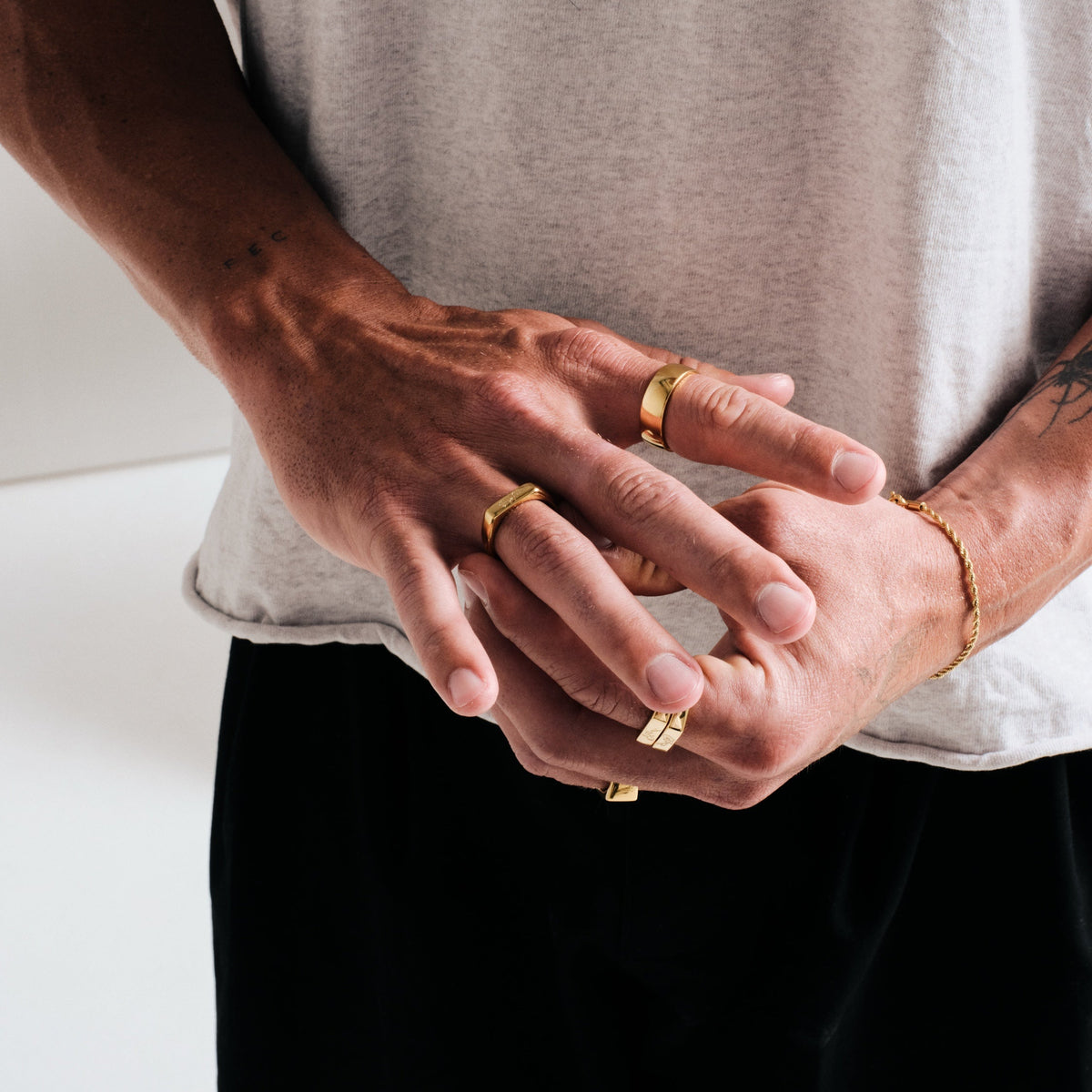 Man wearing gold stainless rings, hands clasped at the waist, with a gold rope bracelet on left wrist