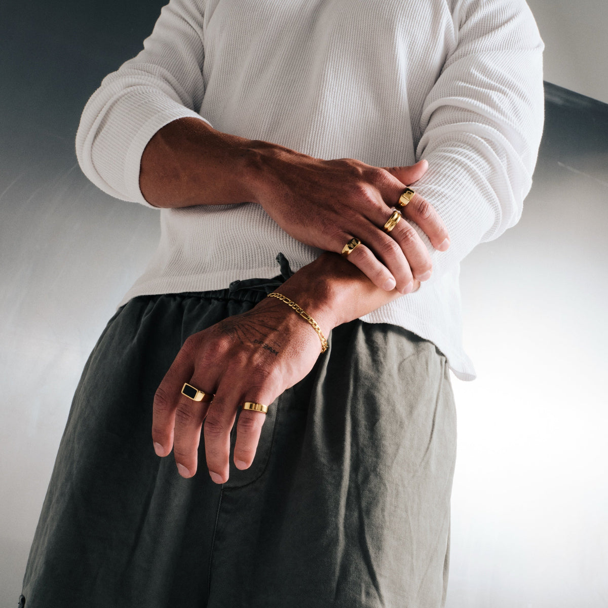 Man wearing gold stainless rings, arms folded, low angle, with a gold Figaro bracelet on left wrist