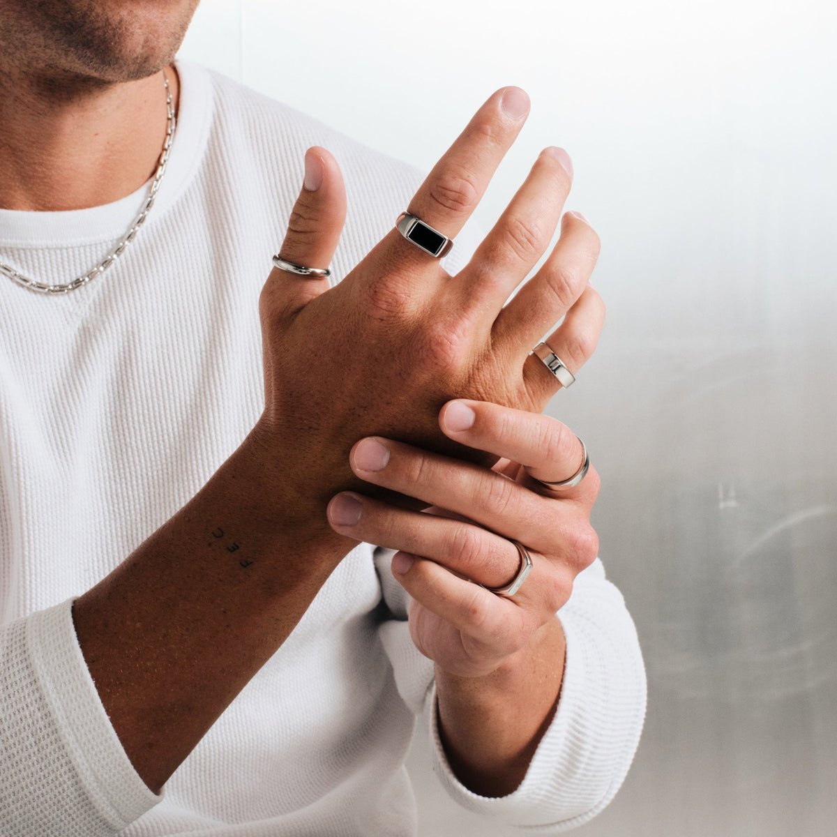 Man wearing silver finish stainless rings, one hand holding the other wrist, with a steel chain necklace