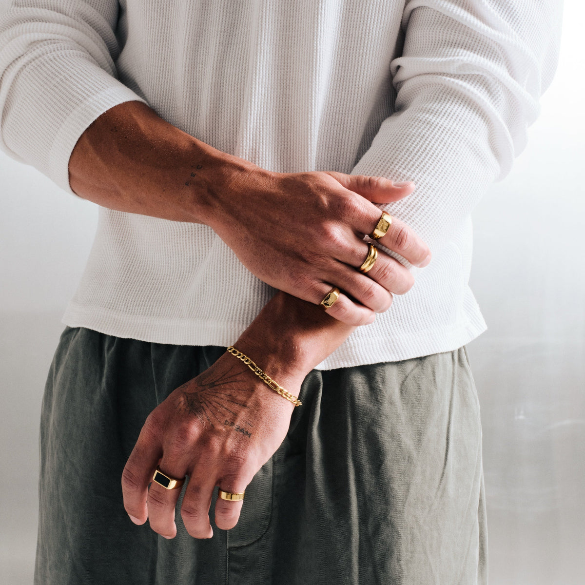 Man wearing gold stainless rings, arms crossed in front, with a gold Figaro chain bracelet, white waffle top, olive pants