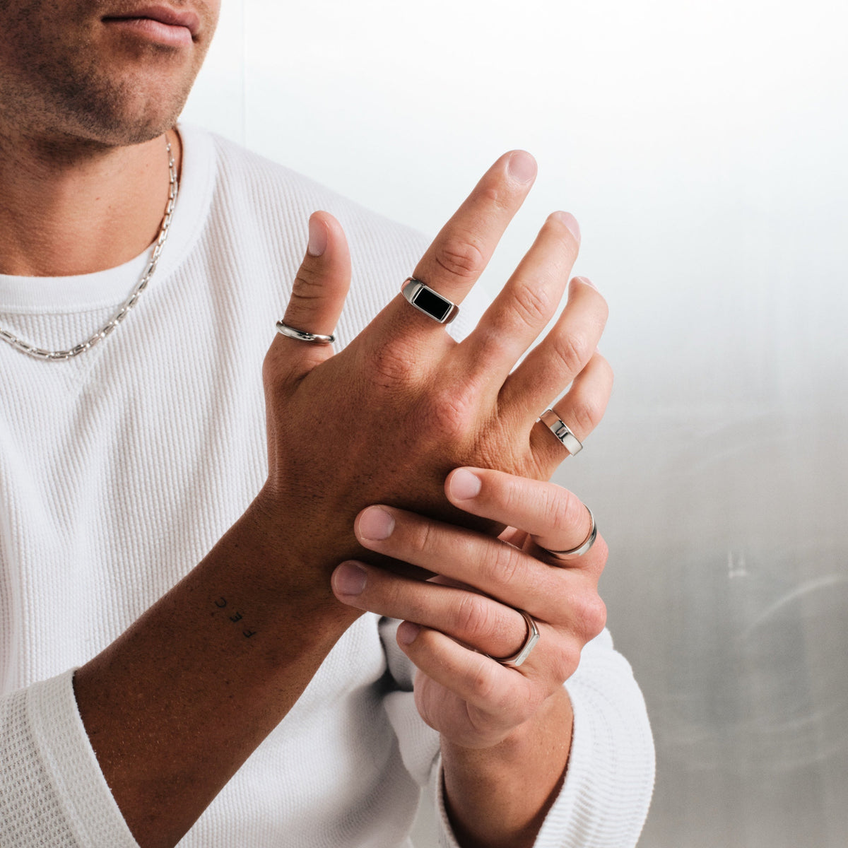 Man wearing silver finish stainless rings, hand raised, other hand holding it, with a steel chain necklace, white waffle top