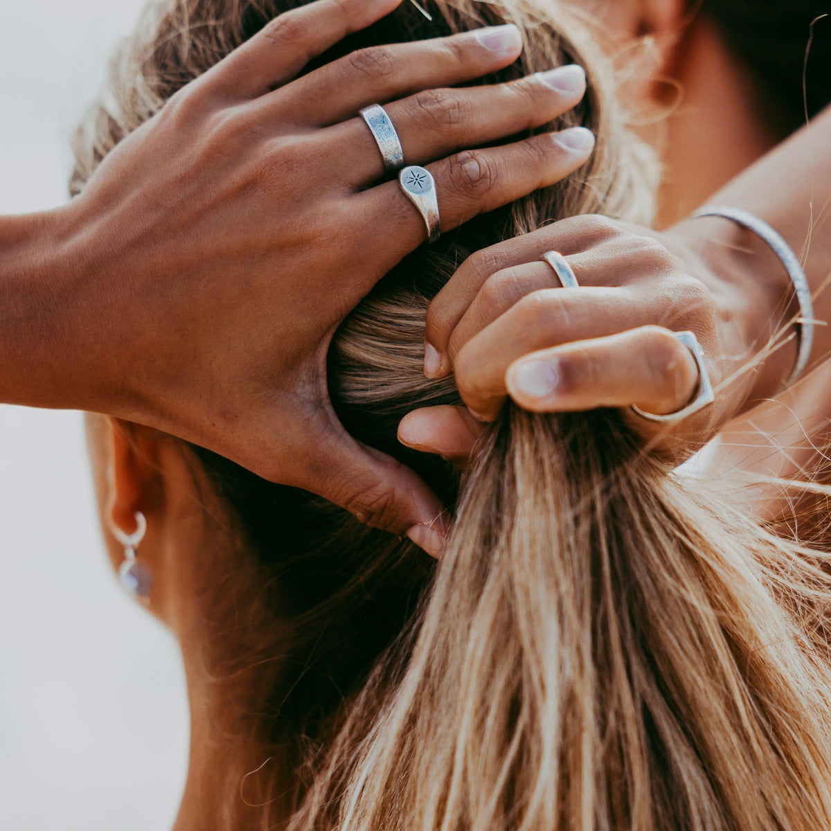 A man's hands wearing the sterling silver Luminous Ring and other Billie Jo jewellery run through a woman's blonde hair in a sunlit, editorial shot.