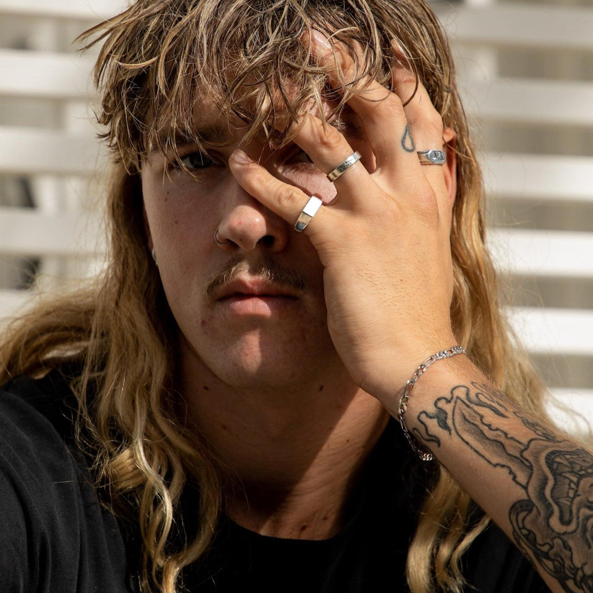 A male model with long blonde hair and a moustache, wearing a stack of sterling silver rings from Billie Jo, including the Genesis Ring. A modern, masculine look from this Australian jewellery brand.