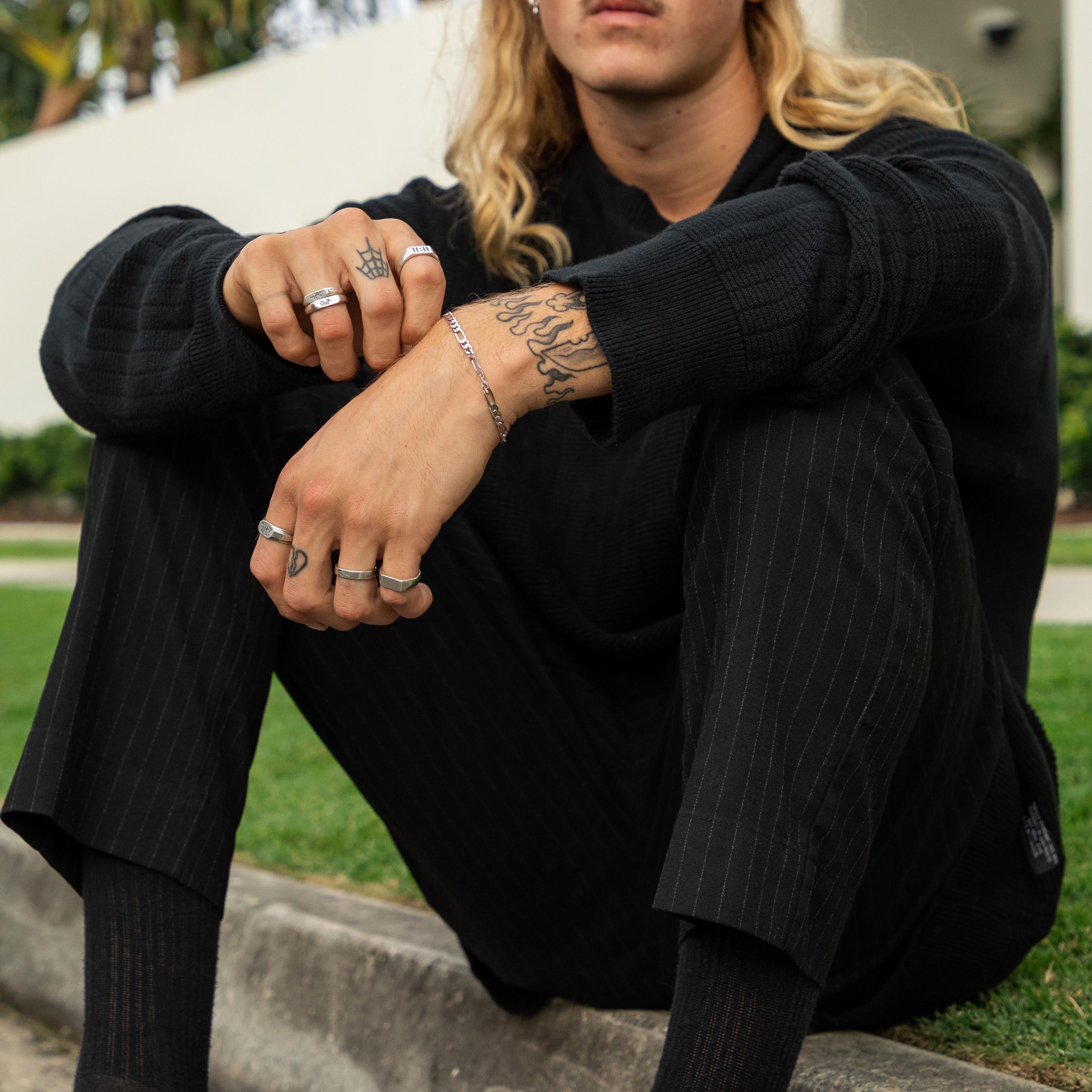 A male model with tattoos wears the Billie Jo Figaro Bracelet with an all-black outfit. This lifestyle shot shows how the sterling silver bracelet adds a point of interest to a dark, modern look.