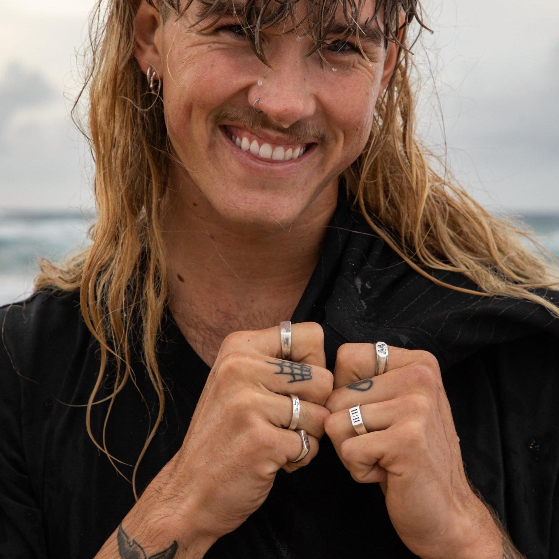 A smiling man with a mullet and nose ring wears a stack of sterling silver rings from Australian brand Billie Jo, including the Prowess Ring, in a cool beachside shot.