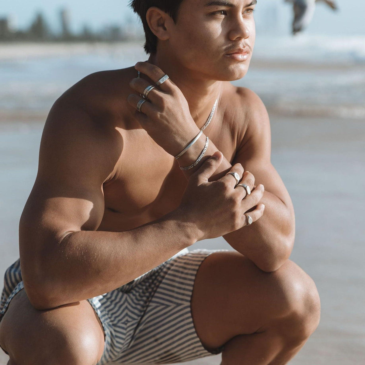 A man crouching on the beach wearing the Billie Jo Mariner Bracelet and other silver jewellery, embodying the brand's modern and masculine aesthetic.