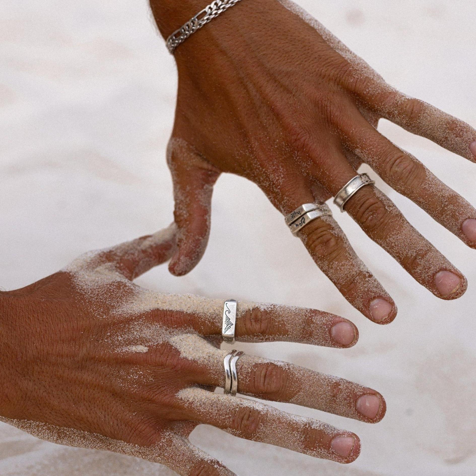 A man's hands, dusted with white sand, wearing a stack of sterling silver rings from Australian men's jewellery brand Billie Jo, including the Union Ring.