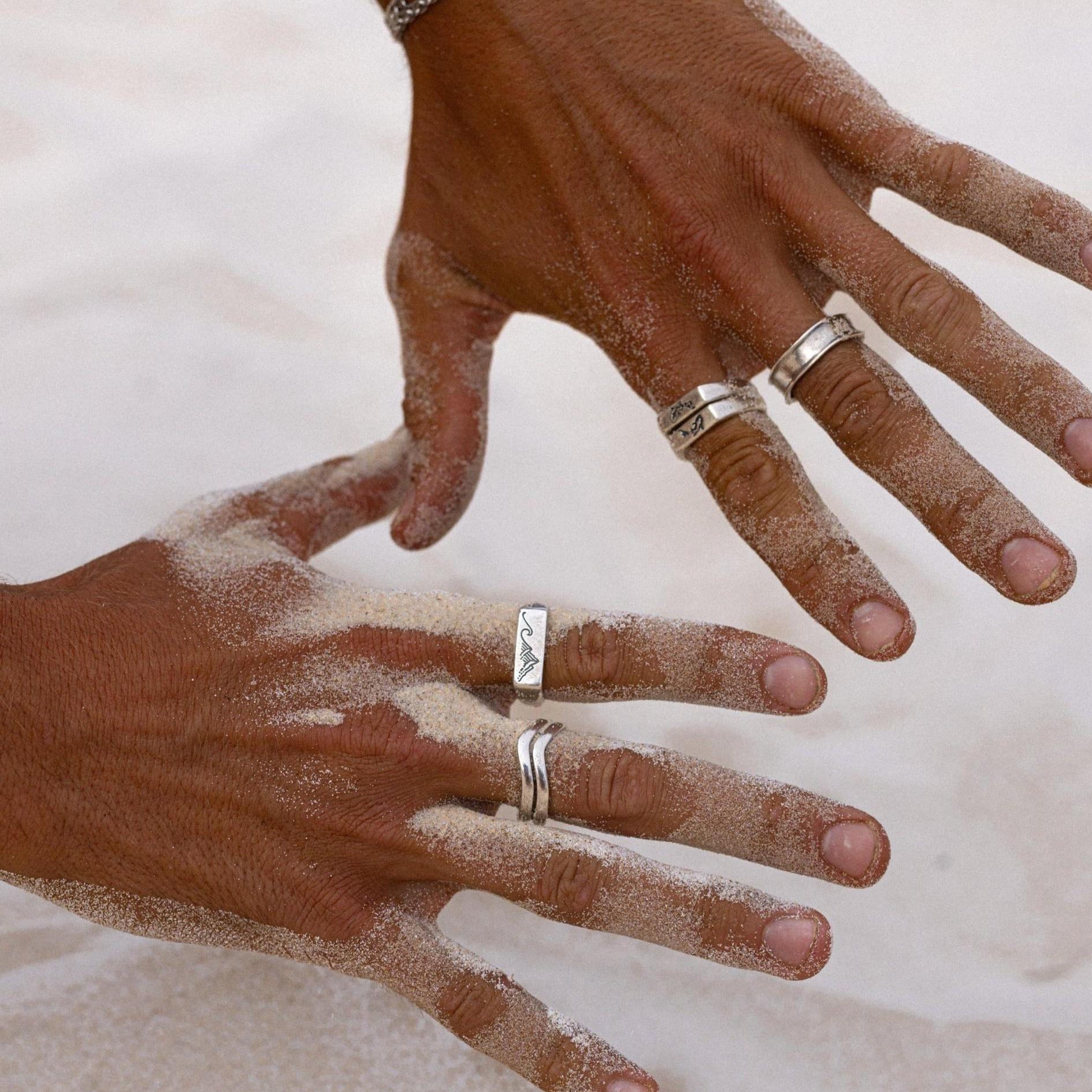 A close-up of a man's hands covered in sand, wearing a collection of sterling silver rings from Australian jewellery brand Billie Jo. The image highlights the texture and detail of the rings in a beach setting.