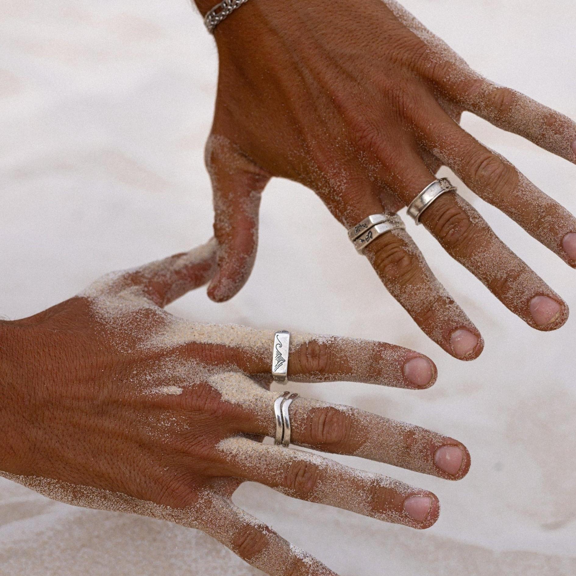 Two hands covered in fine white sand, showcasing a selection of sterling silver rings from Billie Jo, including the distinctively shaped Forge Ring.