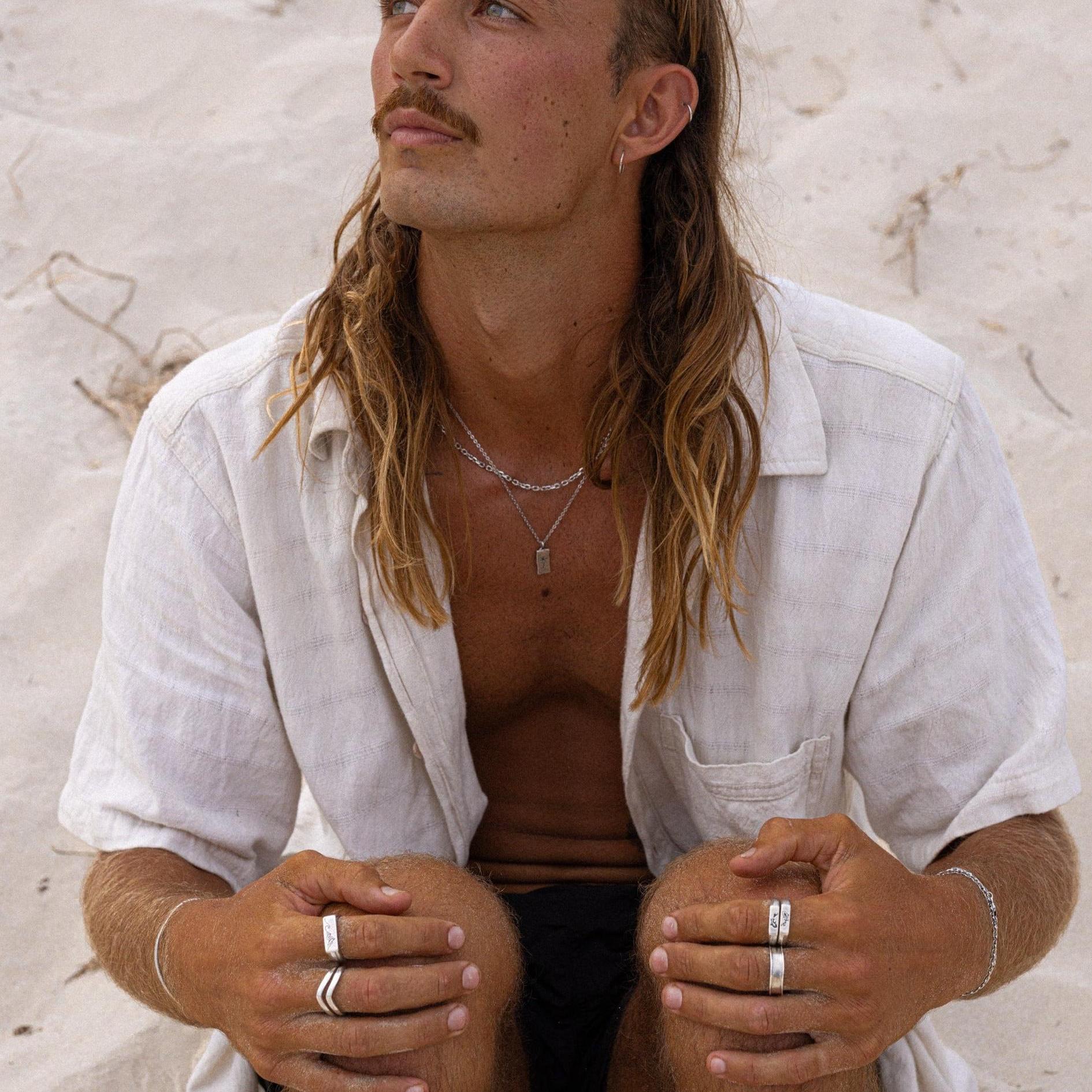 A male model with long hair sits on the sand wearing the sterling silver Paradiso Pendant from Billie Jo, layered with other silver necklaces.