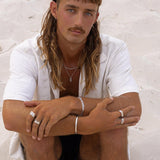 A male model with a mullet and moustache wears the Billie Jo Figaro Bracelet, a silver cuff, and multiple rings. This editorial shot for the Australian men’s jewellery brand has a modern, beachy aesthetic.