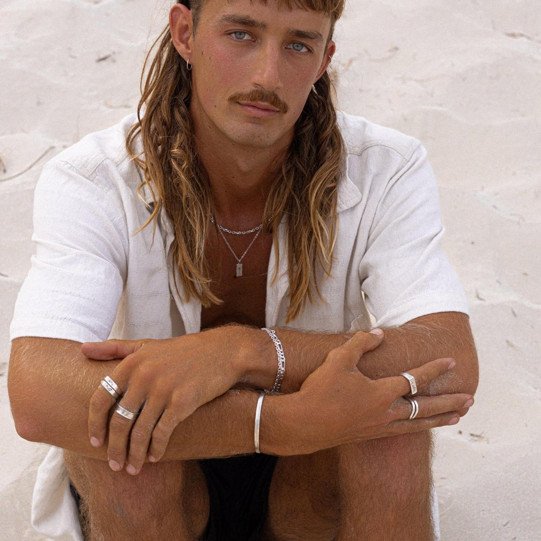 A male model with a mullet and moustache wears the Billie Jo Figaro Bracelet, a silver cuff, and multiple rings. This editorial shot for the Australian men’s jewellery brand has a modern, beachy aesthetic.