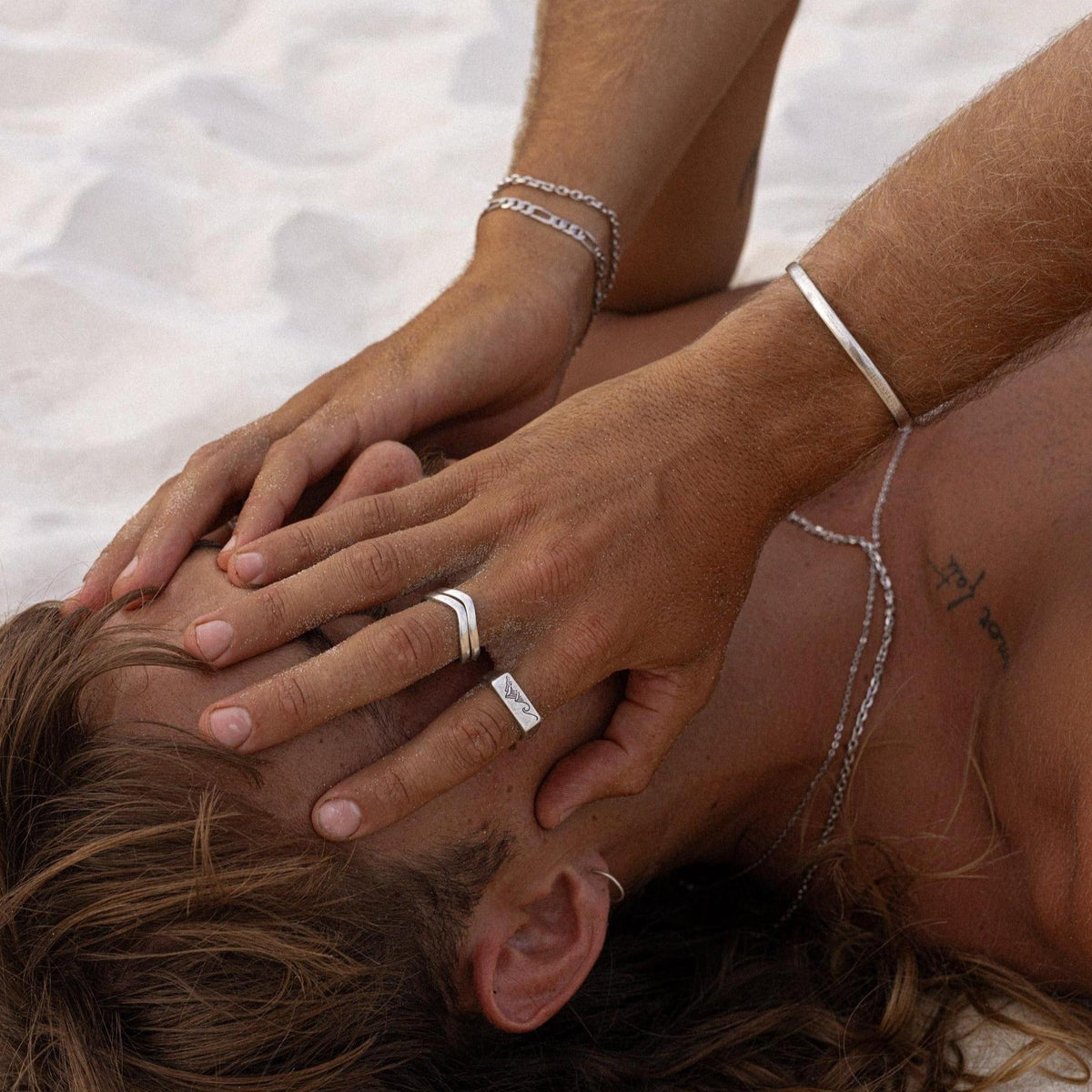 A man lies on the sand, his hands adorned with the sterling silver Batch Bracelet and other jewellery from Billie Jo. A relaxed, editorial shot showcasing Australian men's style.