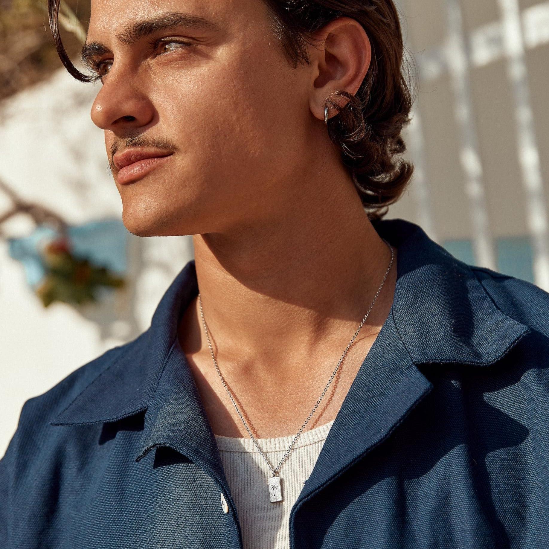 A male model wearing the sterling silver Paradiso Pendant from Billie Jo, styled with a white tank top and an open blue shirt for a casual look.