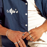 A stylish man wearing a collection of Billie Jo men's jewellery, including the engraved Essence Ring, paired with a casual blue button-up shirt.