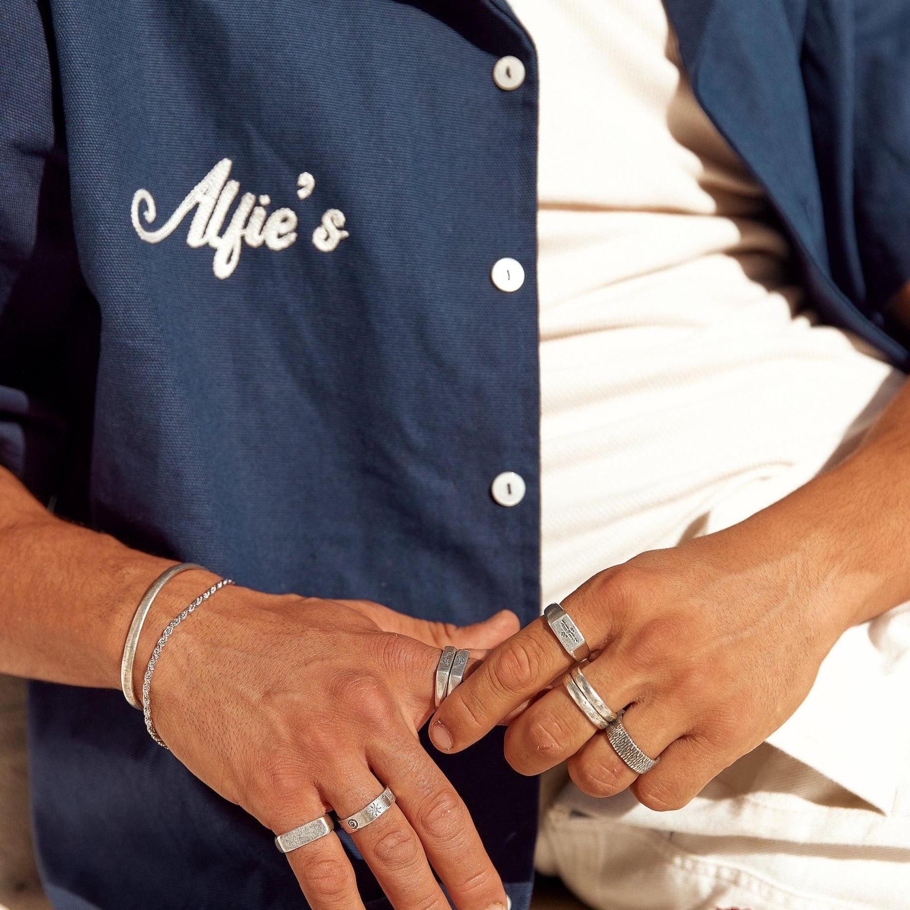 A stylish man wearing a collection of Billie Jo men's jewellery, including the engraved Essence Ring, paired with a casual blue button-up shirt.
