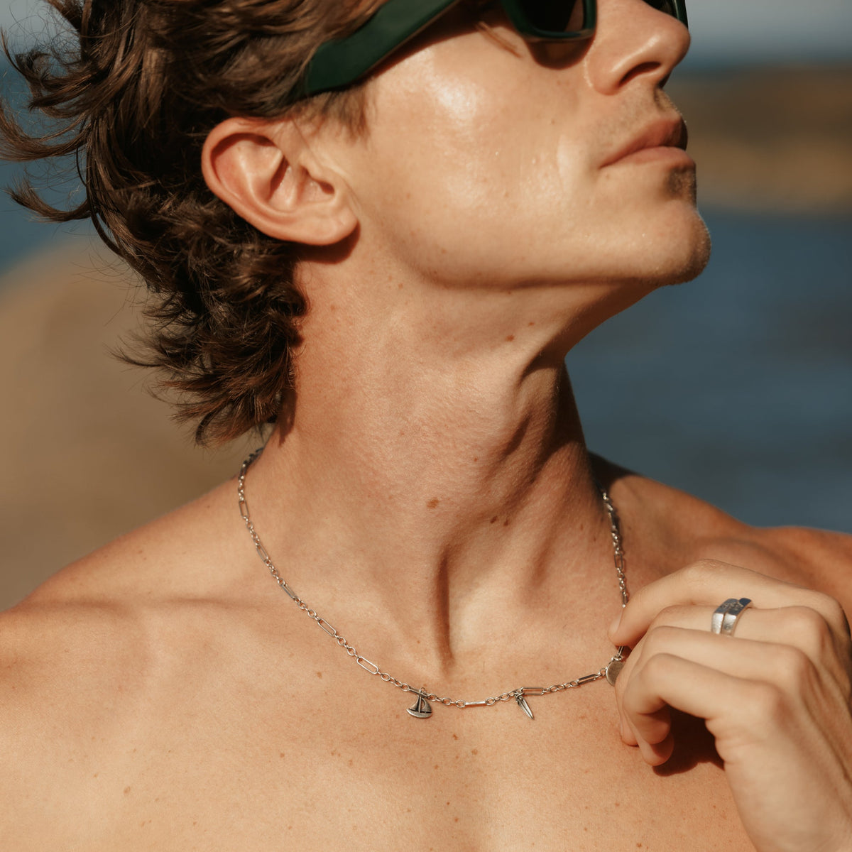 A man wearing sunglasses adjusts his sterling silver Freedom Necklace from Billie Jo, with the ocean in the background, capturing a Gold Coast summer aesthetic.