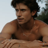 A male model wears the Billie Jo Freedom Necklace, showcasing its sterling silver paperclip chain and unique pendants against his skin in a relaxed, outdoor setting.